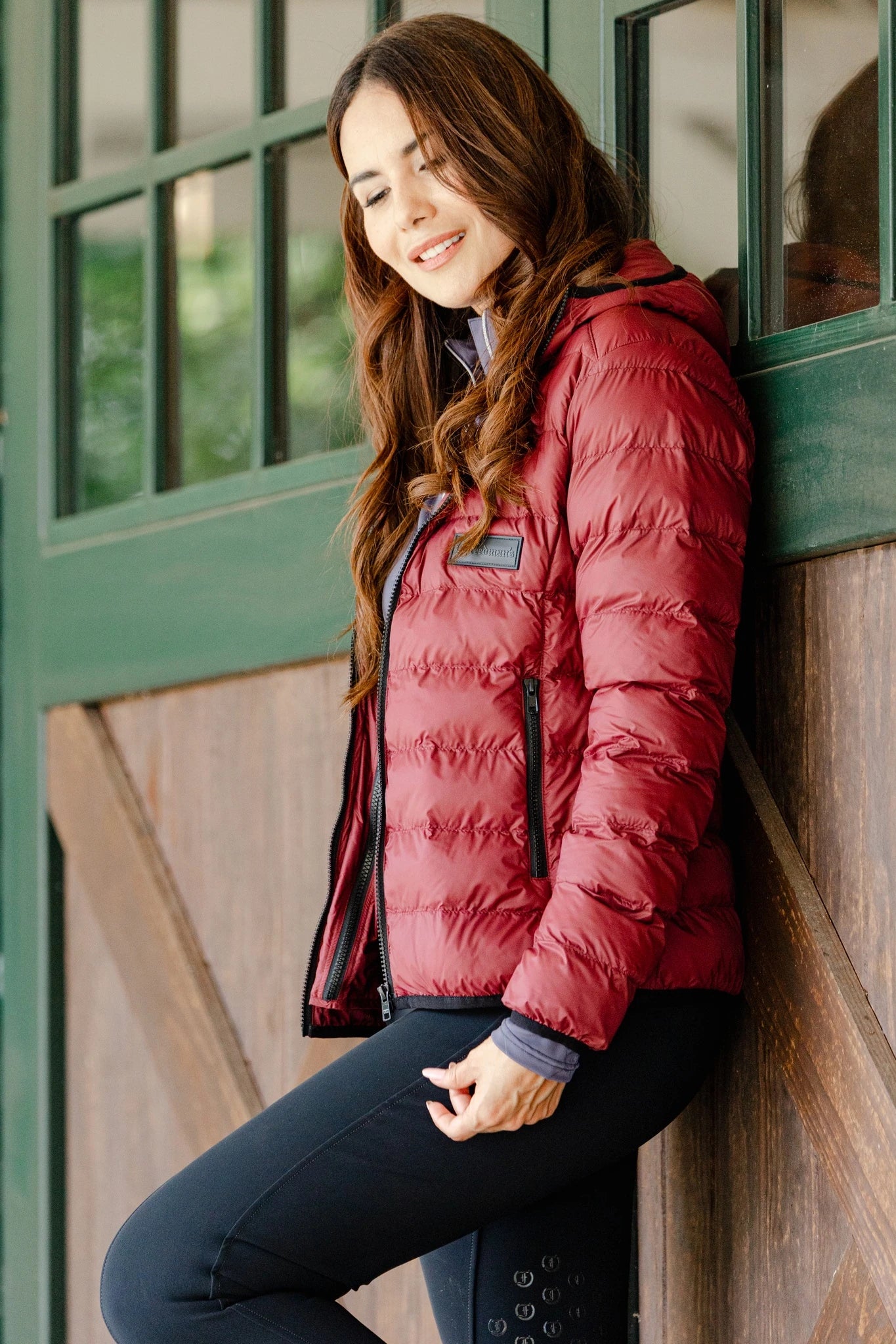 Woman wearing the Signature Ladies' Packable Puffer Jacket in burgundy, featuring a quilted design, front zipper, zippered pockets, and hood, leaning against a stable door.