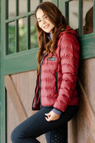 Woman wearing the Signature Ladies' Packable Puffer Jacket in burgundy, featuring a quilted design, front zipper, zippered pockets, and hood, leaning against a stable door.