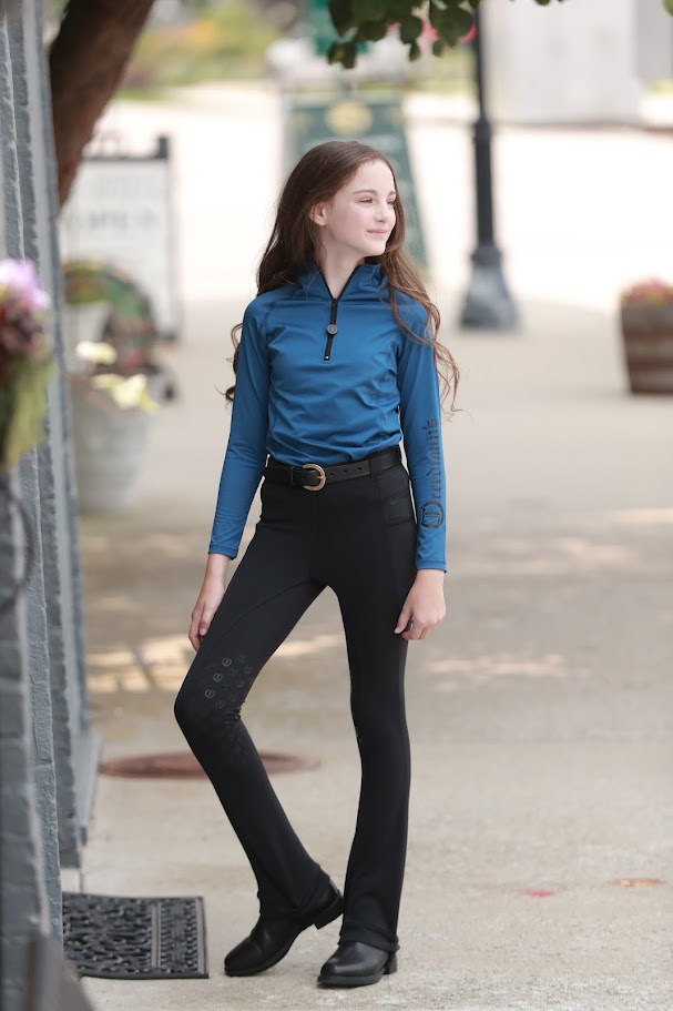 Young girl wearing the Freedman's Kids 1/4 Zip Base Layer in blue, paired with black riding breeches, a belt, and black boots. She stands confidently outdoors, showcasing the stylish and functional equestrian top with a sleek zip-up design and Freedman’s logo on the sleeve. Perfect for riding and active wear.