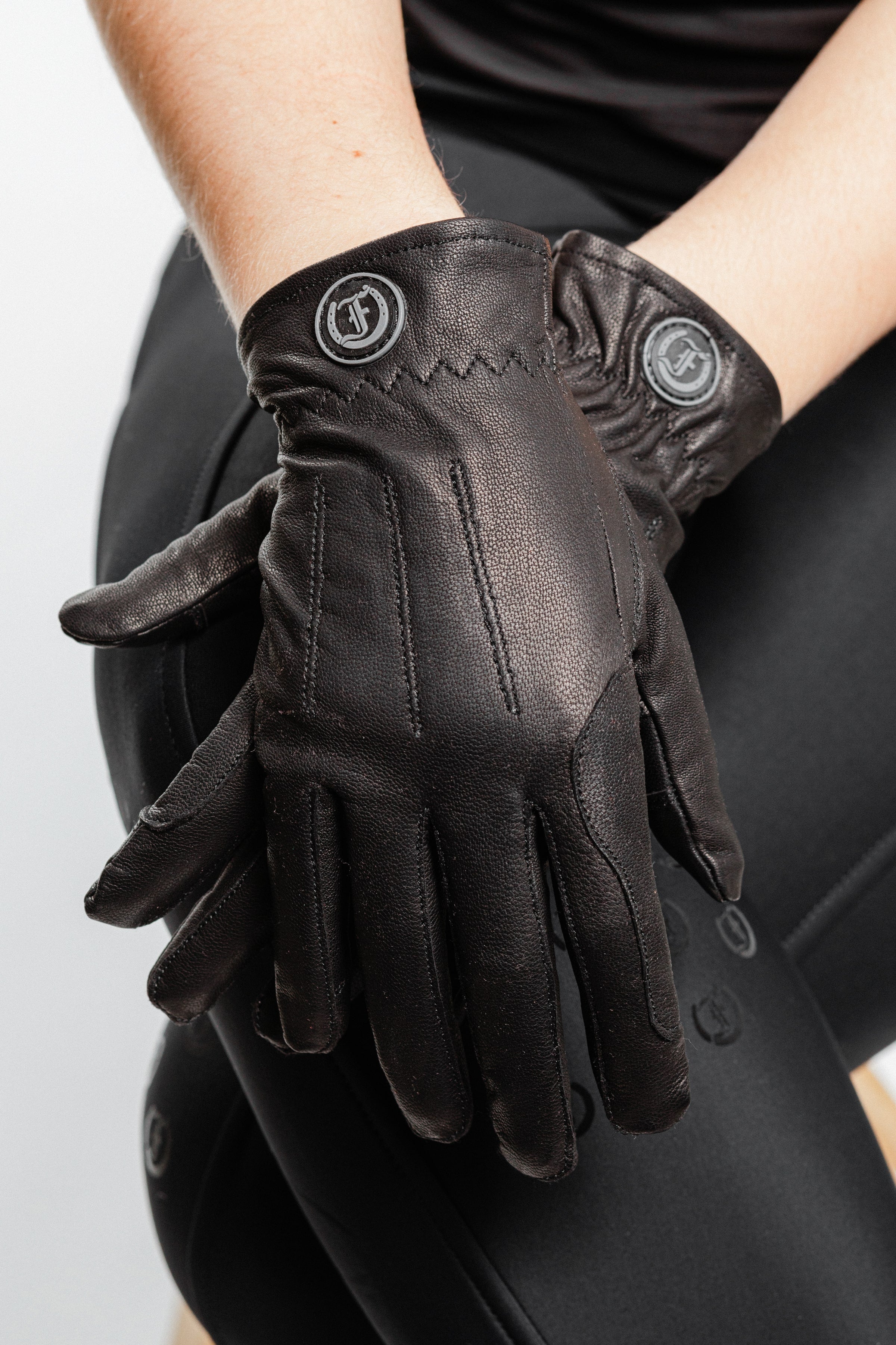 Tous les Jours Gloves, dark gray leather, worn on two hands with embossed circular logo on wrist straps, visible seam stitching and textured surface, dark clothing visible in background.