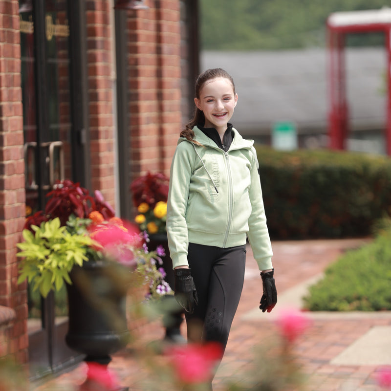 Young girl wearing the Freedman's Kid's Green Logo Hoodie in mint green, paired with black riding leggings and gloves. She is walking outdoors near a brick building with flowers, smiling confidently. The hoodie features a zip-up design and an embroidered Freedman’s logo, perfect for equestrian and casual wear.