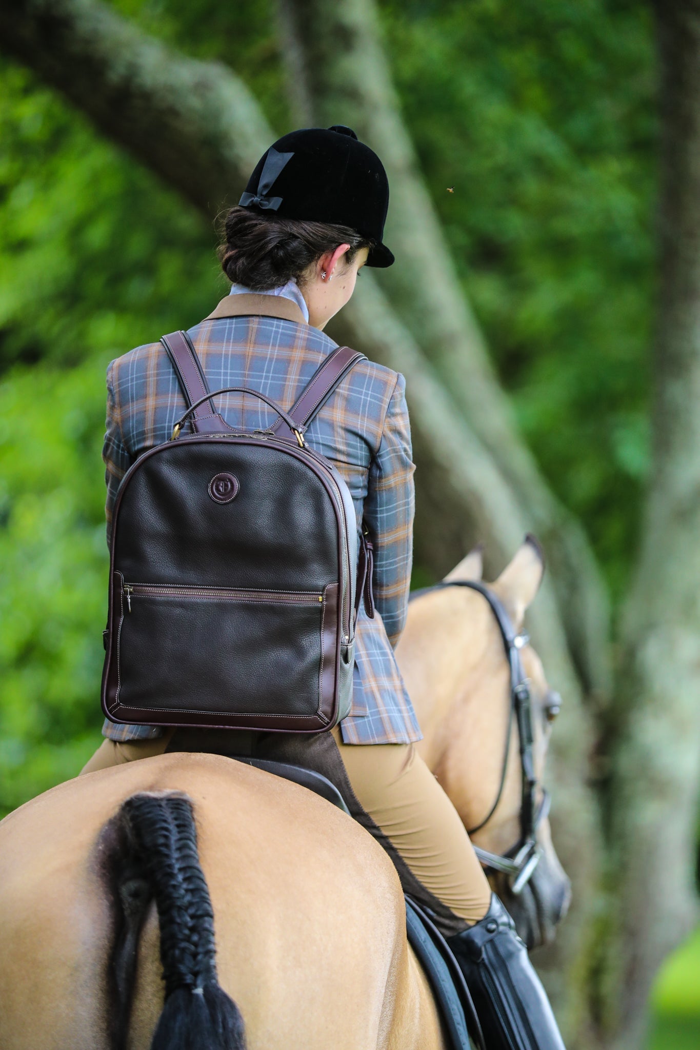 Equestrian rider wearing a plaid jacket and black velvet helmet, carrying 'The Jaunty Backpack' by Freedman—a sophisticated brown leather backpack with gold accents—while riding a beautifully groomed horse through a lush green setting