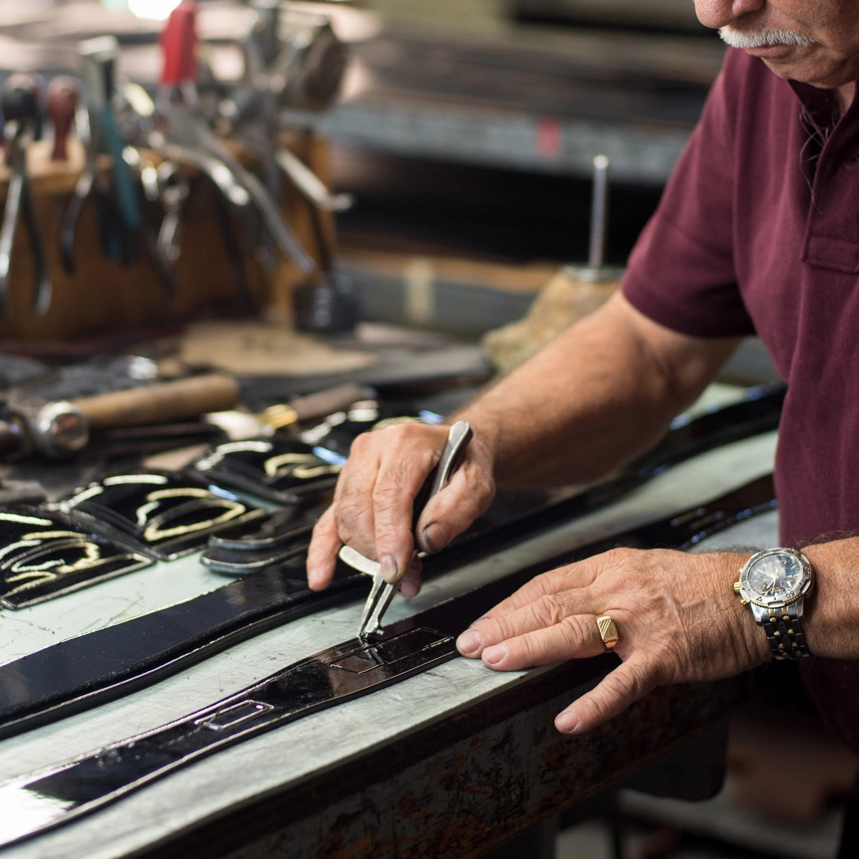 A close-up of a craftsman's hands marking a black leather strap on a workbench with a metal ruler, in a workshop setting.