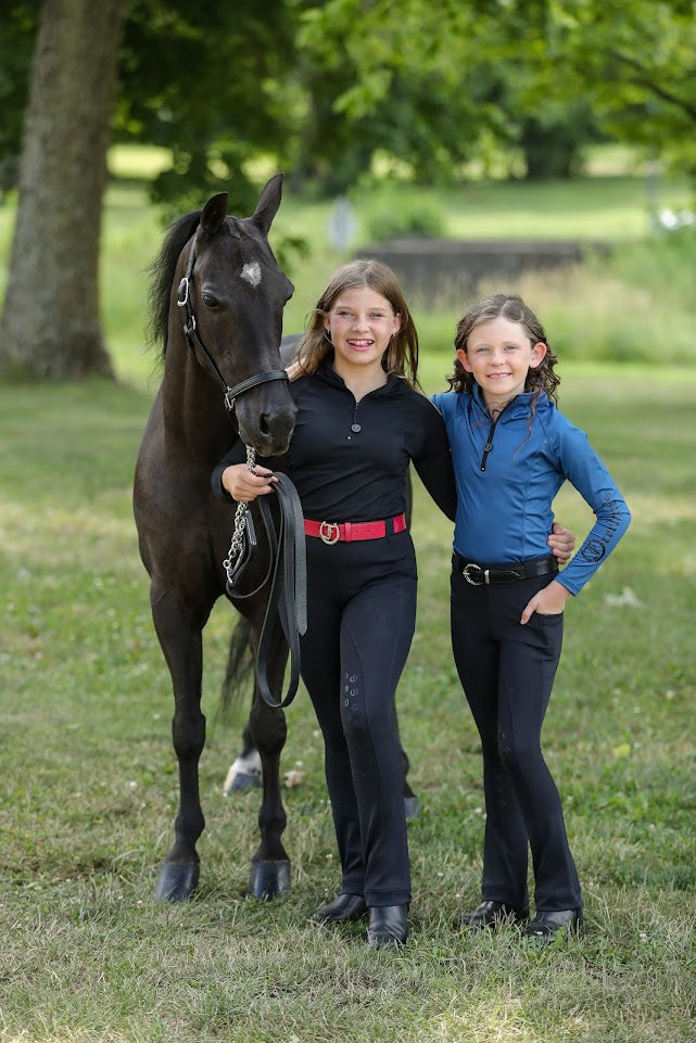 Two Young girls wearing the Freedman's Kids 1/4 Zip Base Layer one in blue and other in black, paired with black riding breeches, a belt, and black boots. She stands confidently outdoors, showcasing the stylish and functional equestrian top with a sleek zip-up design and Freedman’s logo on the sleeve. Perfect for riding and active wear.