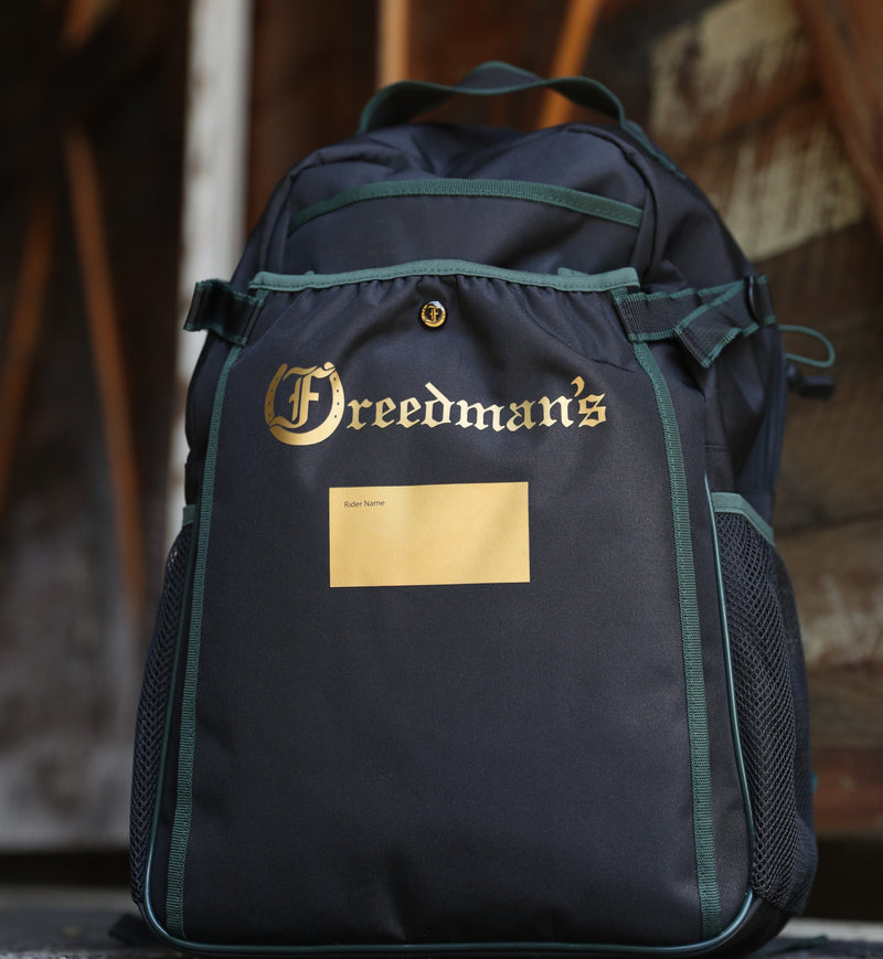 Team Freedman's Backpack, black fabric with green trim, gold Freedman's logo, gold rider name slot, mesh side pockets, and adjustable straps, outdoor setting.