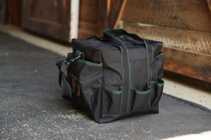 Team Freedman's Grooming Bag, black fabric with green trim, multiple exterior pockets, green webbing handles, visible buckle strap, and outdoor setting.