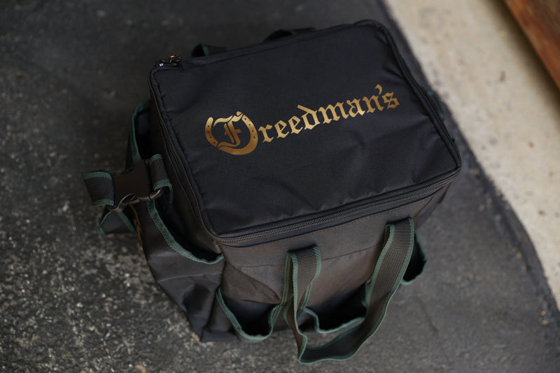 Team Freedman's Grooming Bag, black fabric with gold Freedman's logo, green webbing handles, multiple zippered compartments, and outdoor setting.