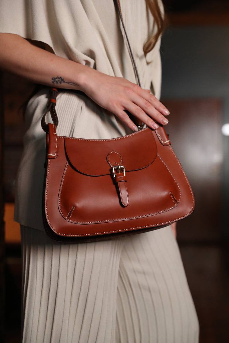 A woman in beige holds a brown leather Buckingham Bag  with contrast stitching and a buckle closure.