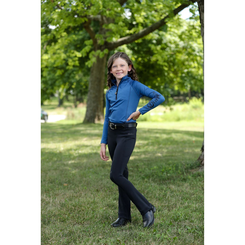 Young girl wearing the Freedman's Kids 1/4 Zip Base Layer in blue, paired with black riding breeches, a belt, and black boots. She stands confidently outdoors, showcasing the stylish and functional equestrian top with a sleek zip-up design and Freedman’s logo on the sleeve. Perfect for riding and active wear.