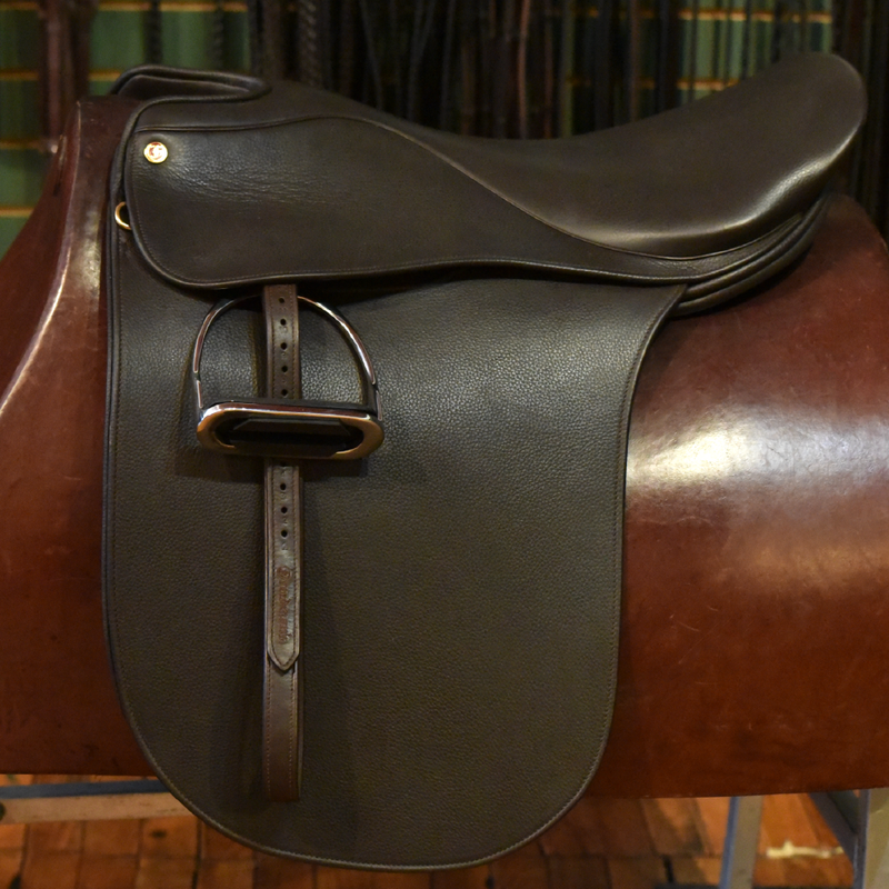 Dark brown leather saddle with attached stirrup leathers and irons, displayed on a saddle stand with green and gold background.