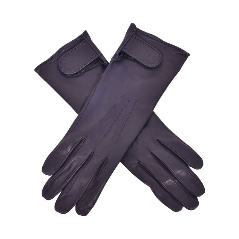 The Yorkville Show Glove, dark navy leather gloves crossed, with wrist strap and button detail, visible seam stitching, light background.
