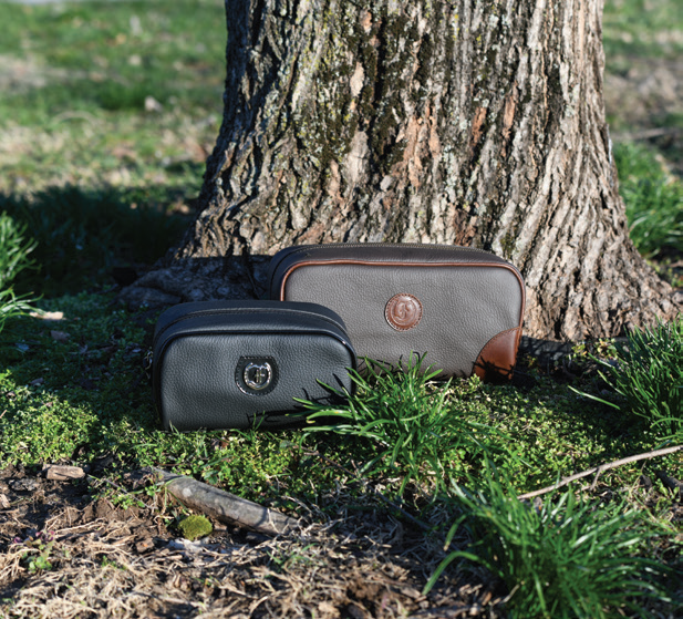 Two Freedman's Hansom Shave Bags, one black leather, one dark brown and tan two-tone leather, embossed "F" logos visible, placed on the ground near a tree trunk and grass, outdoor setting